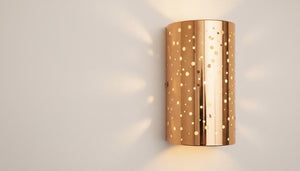 A modern cylindrical wall sconce with a metallic rose gold finish and a pattern of small circular cutouts, casting a warm yellow light onto a neutral beige wall.