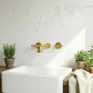 A matte brass wall-mounted faucet and matching side lever on a white plaster bathroom wall above a white apron-front sink, with small potted green plants and a folded beige towel nearby.