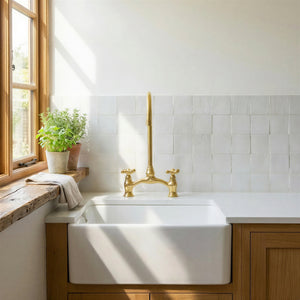 A white farmhouse-style kitchen sink with a tall polished brass faucet and two separate hot and cold handles, installed in a wooden cabinet beneath a white tiled backsplash. A sunlit window with a potted plant and folded towel is visible on the left.