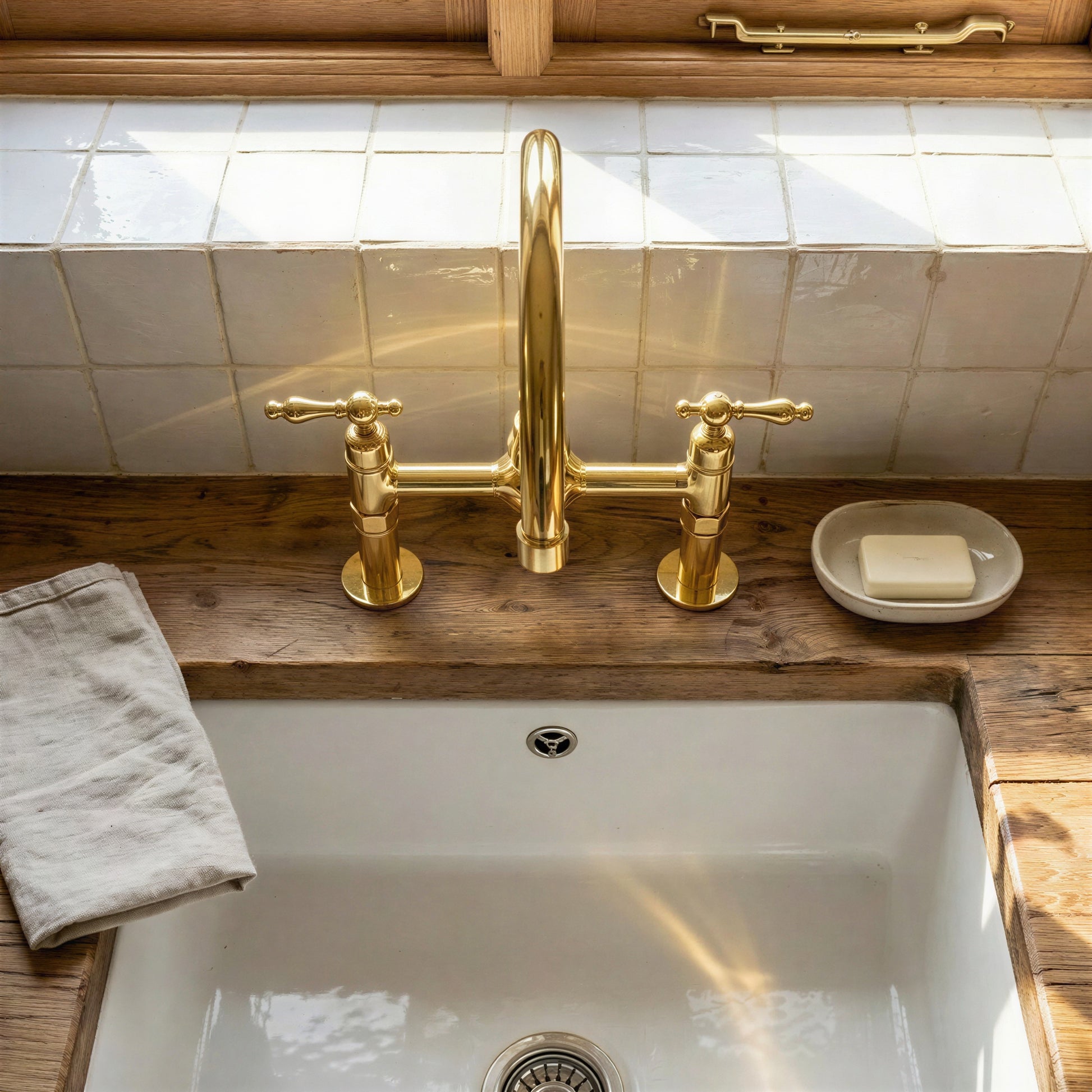 Handmade unlacquered brass bridge kitchen faucet with high-arc gooseneck spout and classic lever handles mounted on a rustic wood countertop over a fireclay sink.