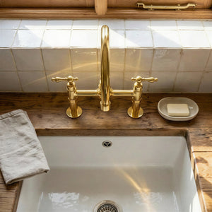 Handmade unlacquered brass bridge kitchen faucet with high-arc gooseneck spout and classic lever handles mounted on a rustic wood countertop over a fireclay sink.