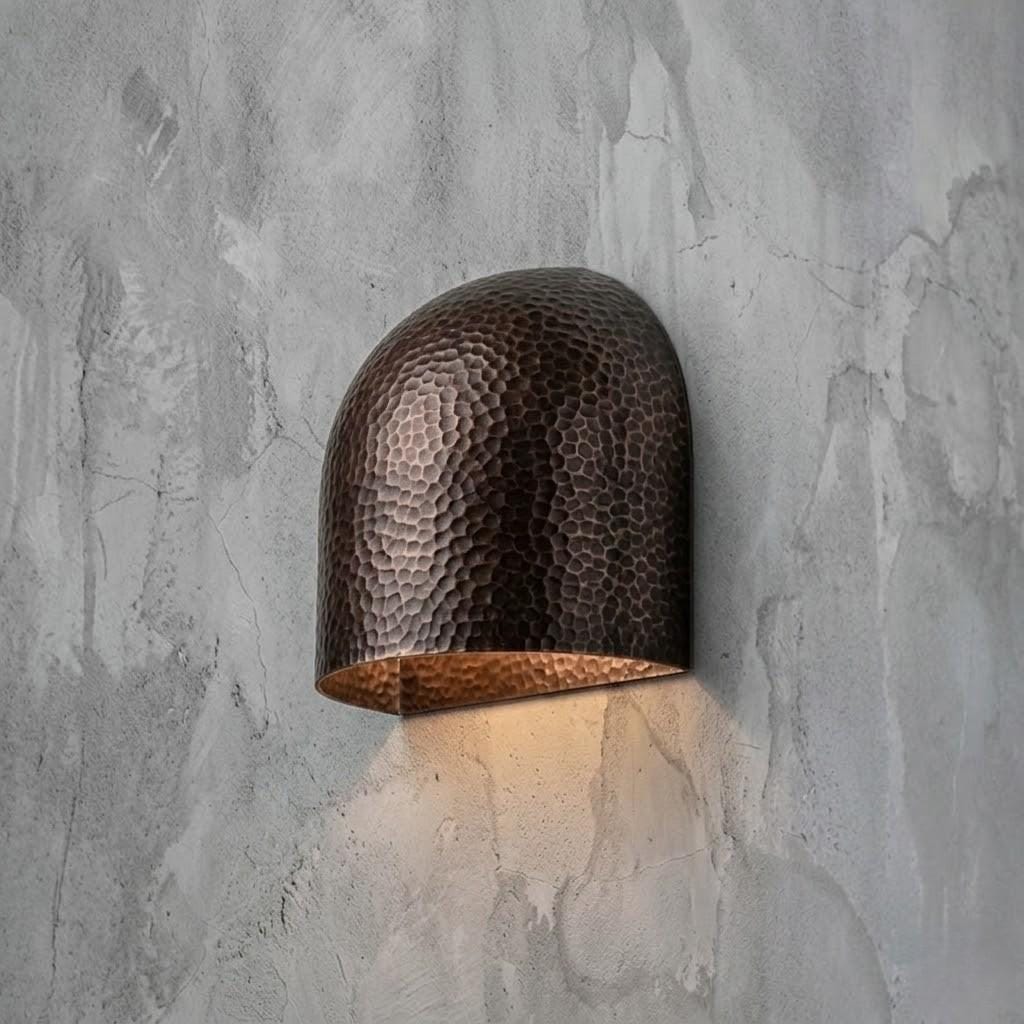 A hammered metal dome wall sconce with a warm copper-toned interior, mounted on a textured gray concrete wall and casting a soft downward light.