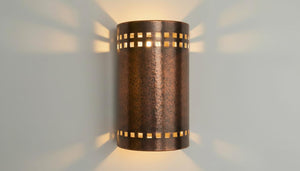 A cylindrical wall sconce with a hammered metallic finish, featuring square cutout patterns near the top and bottom, casting warm light and shadow on a light gray wall.