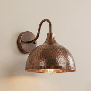 A rustic copper wall sconce with a hammered dome shade and curved arm, mounted on a beige wall and illuminated.