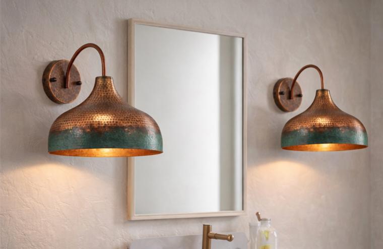 Two matching copper wall sconces with a hammered finish and a green patina lower band, mounted on a textured beige wall on either side of a rectangular mirror.