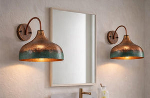 Two matching copper wall sconces with a hammered finish and a green patina lower band, mounted on a textured beige wall on either side of a rectangular mirror.
