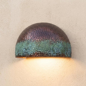A dome-shaped hammered metal wall sconce with a textured finish, featuring a dark bronze top half and a green-blue patina lower half, casting a warm yellow light against a textured beige wall.