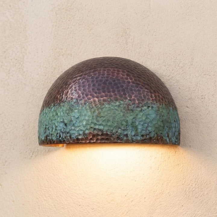 A dome-shaped hammered metal wall sconce with a textured finish, featuring a dark bronze top half and a green-blue patina lower half, casting a warm yellow light against a textured beige wall.