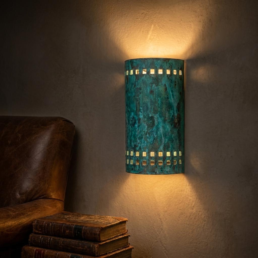 A cylindrical wall sconce with a distressed turquoise-blue metal finish and cutout details, mounted on a textured wall and casting warm light downward, with part of a brown leather chair and stacked books visible at the left.