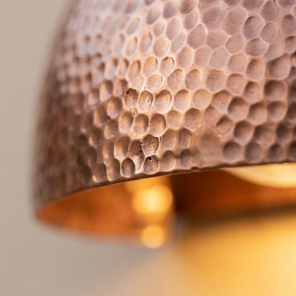 Close-up of a hammered metal dome shade, likely copper or bronze, with a warm glowing light visible inside.