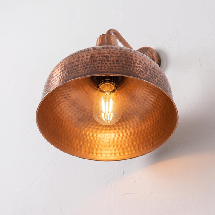 A hammered copper dome wall sconce with a warm glowing light bulb, mounted on a textured white wall.