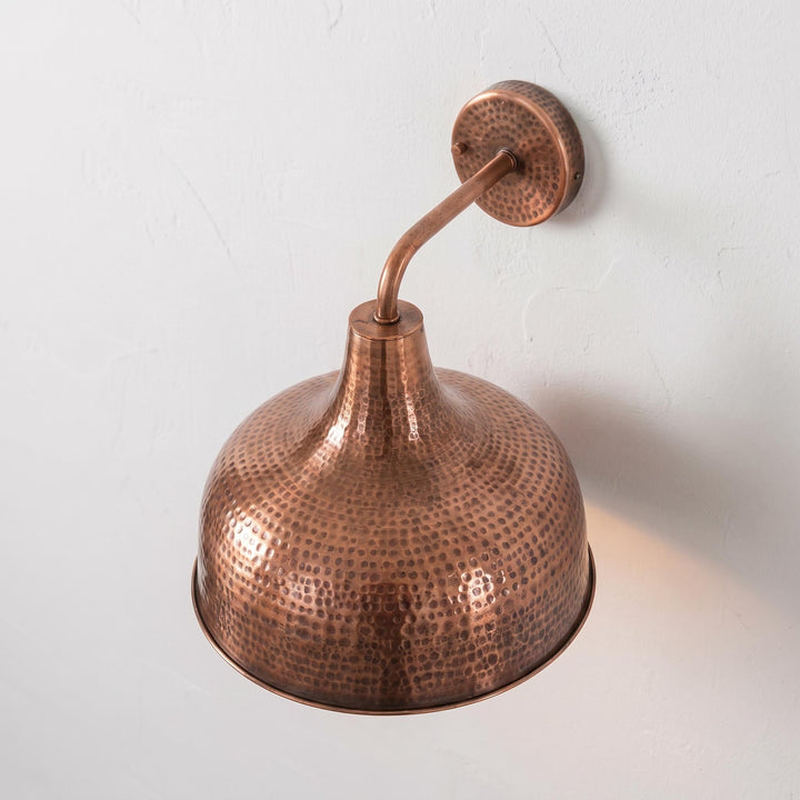 A hammered copper dome wall sconce with a curved arm and round wall mount, shown against a light textured wall.