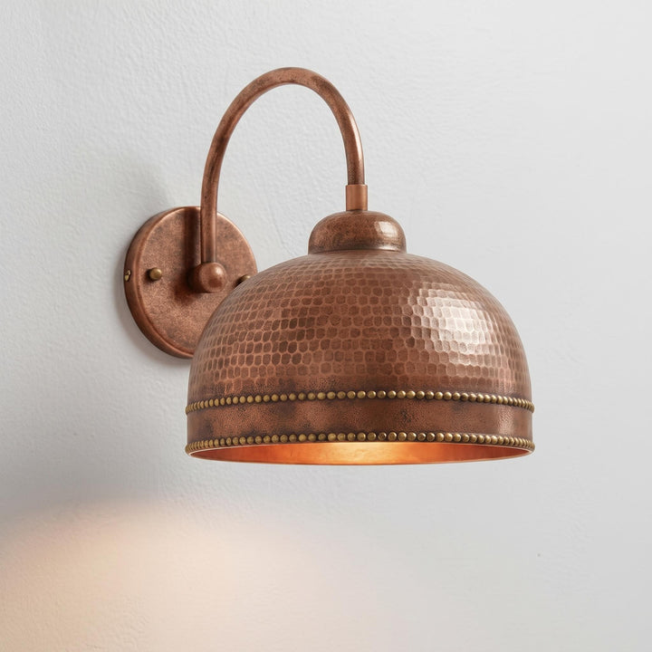 A copper wall sconce with a hammered dome shade and a curved mounting arm, shown lit against a plain light wall.