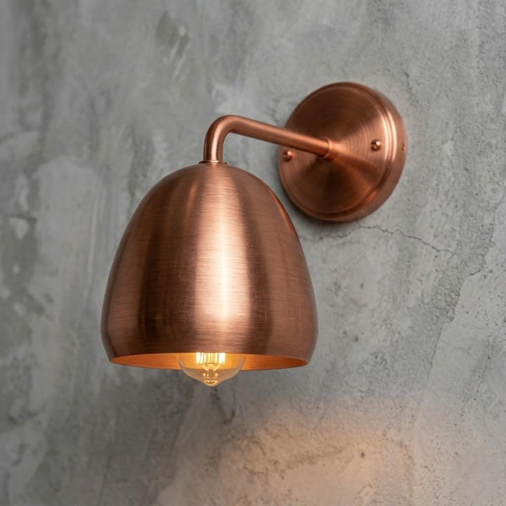 A copper wall sconce with a dome-shaped shade and curved arm, mounted on a textured gray concrete wall and illuminated with a warm light.