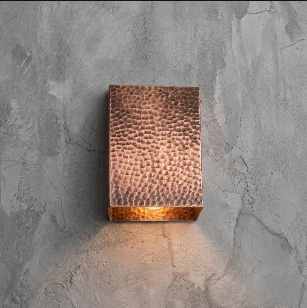 A rectangular hammered copper wall sconce mounted on a textured gray concrete wall, casting a warm light downward.