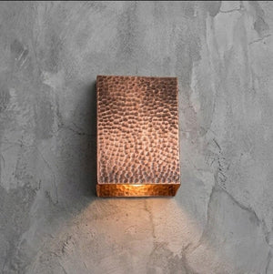 A rectangular hammered copper wall sconce mounted on a textured gray concrete wall, casting a warm light downward.