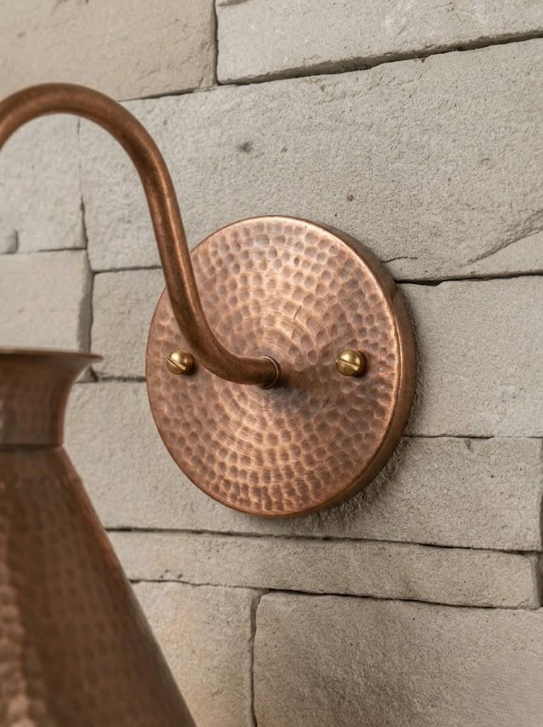 Close-up of a hammered copper wall sconce with a curved hook arm and round circular backplate, mounted on a light stone wall.