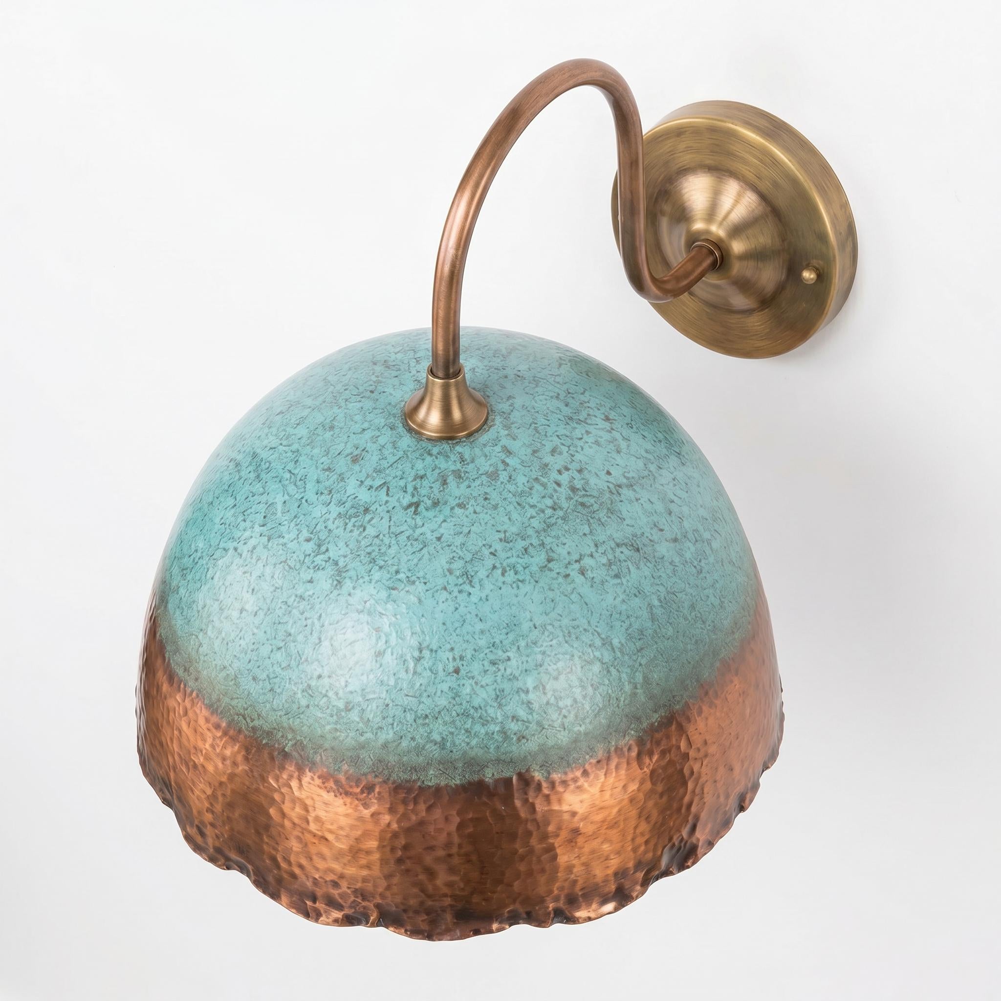 A wall-mounted sconce with a dome-shaped shade featuring a turquoise glaze on the upper half and a hammered copper finish on the lower half, attached to a curved brass arm and round wall plate against a white background.