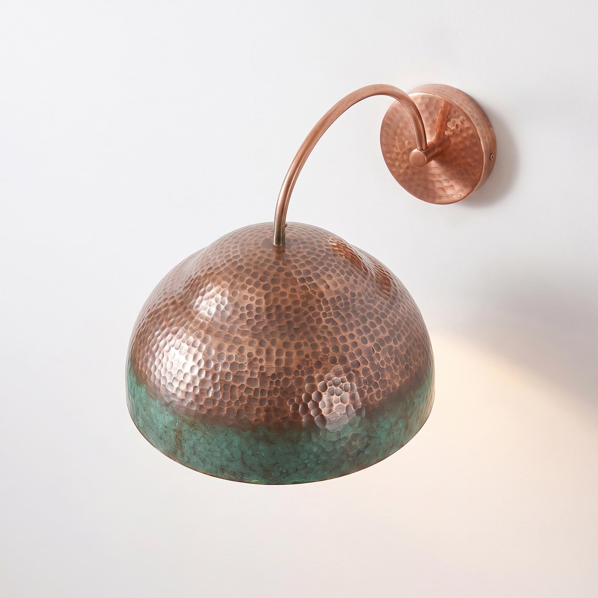 A hammered copper dome wall sconce with a green patina lower edge, mounted on a plain white wall with a warm light shining from the right side.
