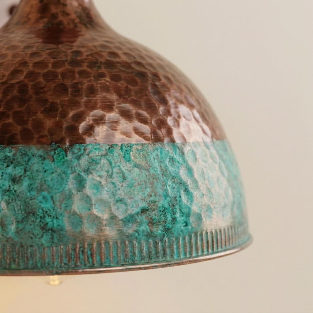 Close-up of a hammered metal dome shade with a split finish: dark brown on the upper half and turquoise patina on the lower half, against a plain light background.