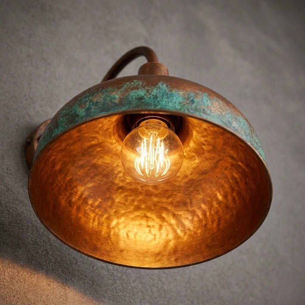 A rustic industrial wall sconce featuring a hammered copper-toned metal shade with a distressed turquoise-green patina rim, housing a glowing warm Edison bulb, mounted on a textured gray wall.