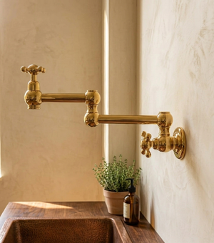 A polished brass wall-mounted kitchen pot filler faucet with cross handles, installed on a textured beige wall above a dark wood sink, with a small potted plant and bottle nearby.