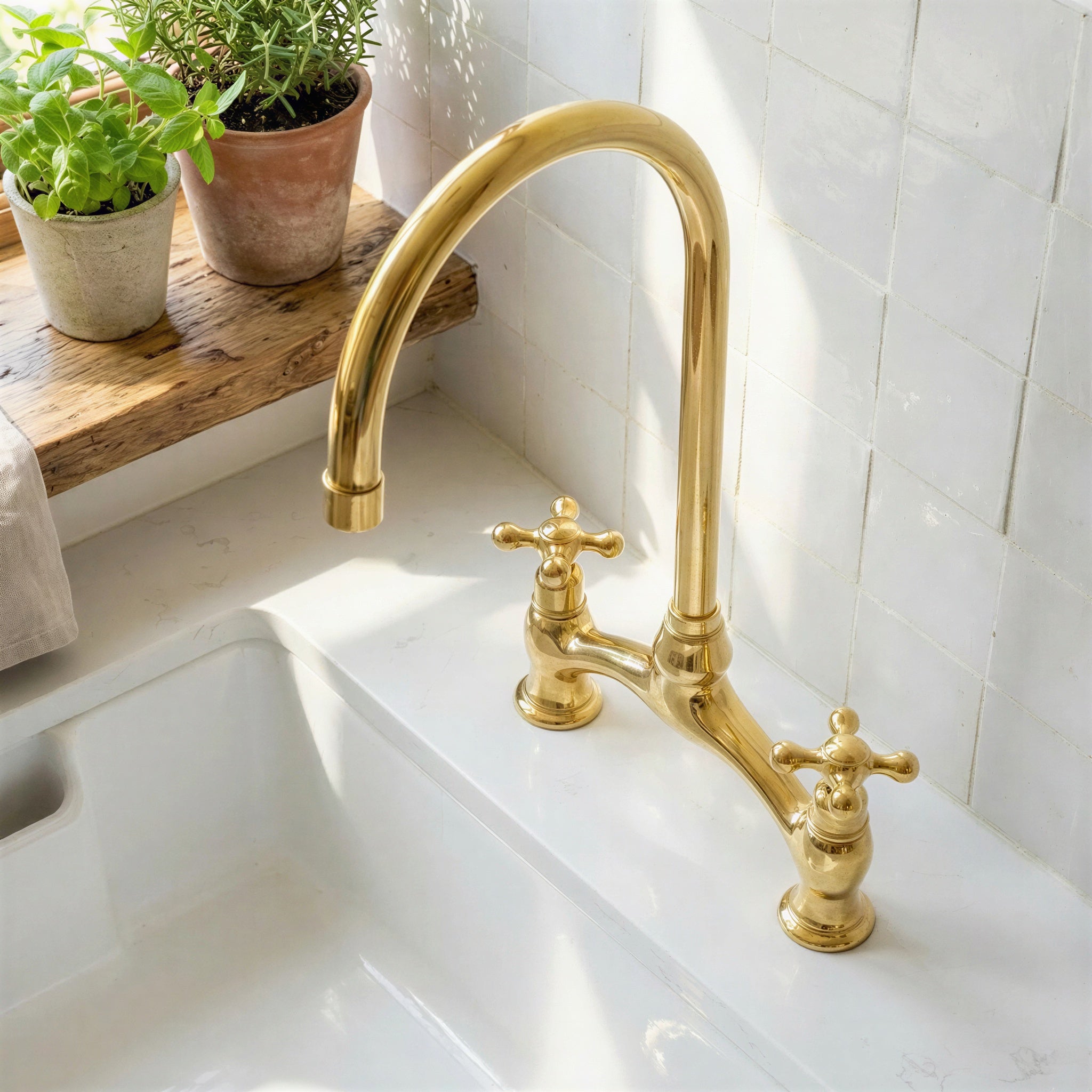 A polished brass bridge faucet with a high-arc spout and two cross handles, installed on a white ceramic kitchen sink with white subway tiles and potted plants in the background.