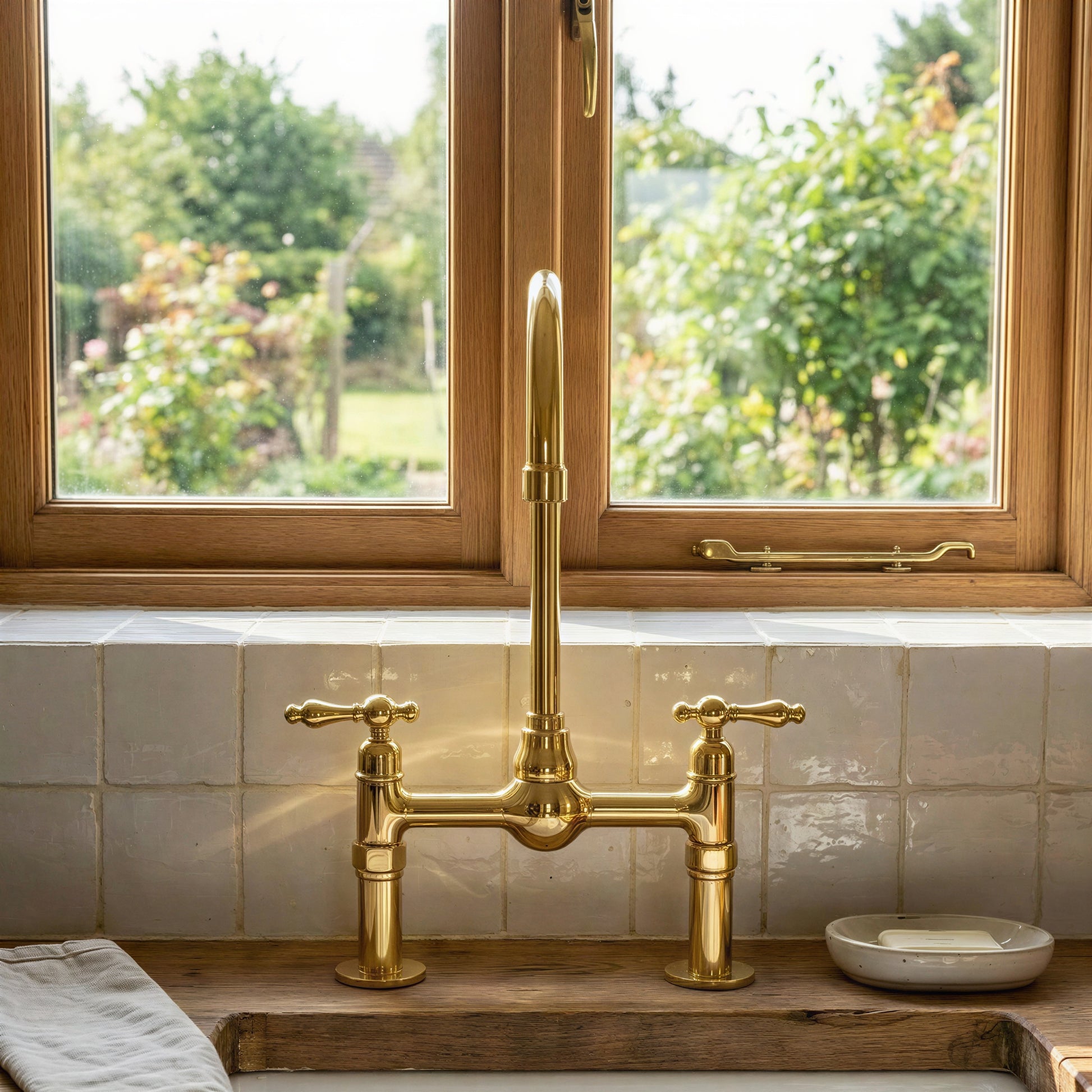 Handmade unlacquered brass bridge kitchen faucet with high-arc gooseneck spout and classic lever handles mounted on a rustic wood countertop over a fireclay sink.