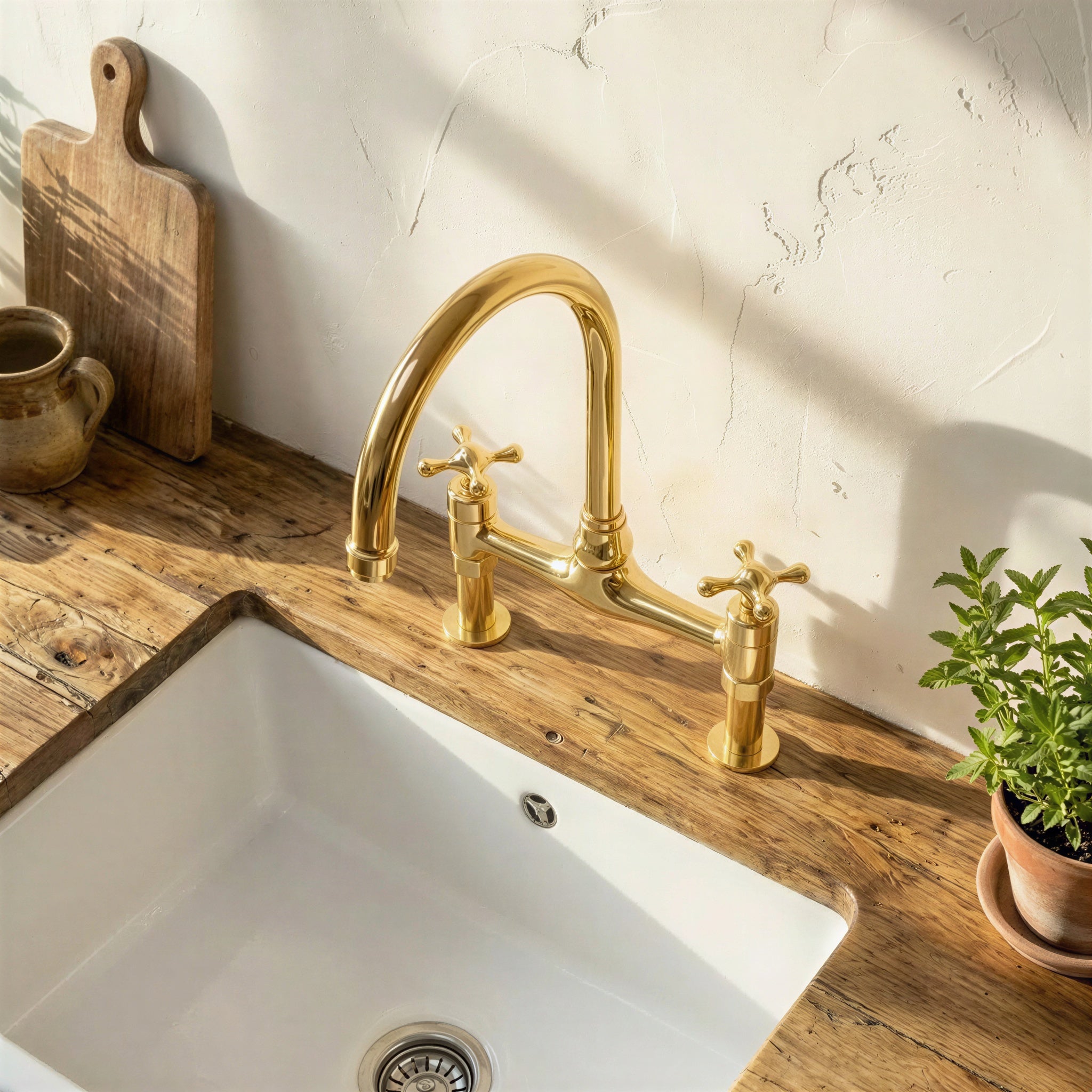 A polished brass bridge faucet with two cross handles installed above a white sink on a rustic wooden countertop, with a white textured wall behind and small decorative props nearby.