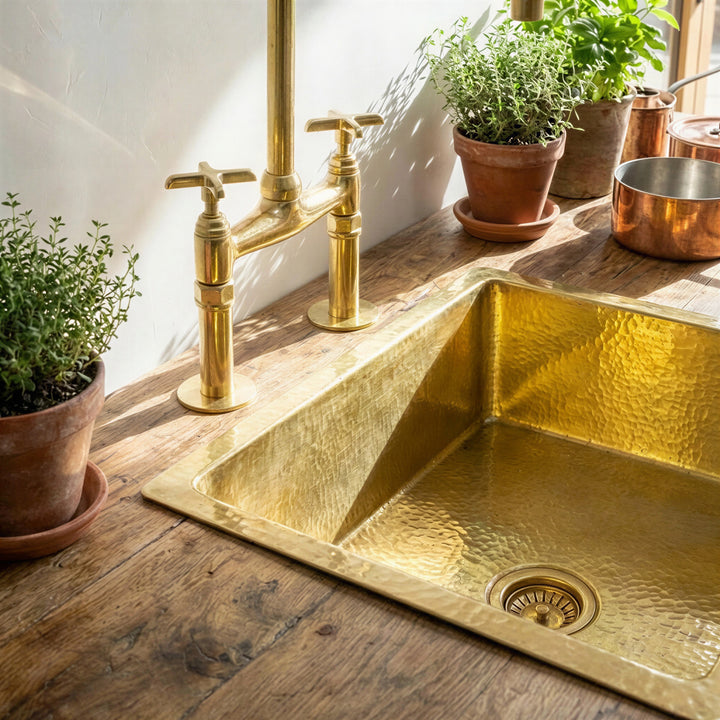 A hammered brass kitchen sink with a matching high-arc bridge faucet set on a rustic wooden countertop, surrounded by potted green plants and warm natural light.