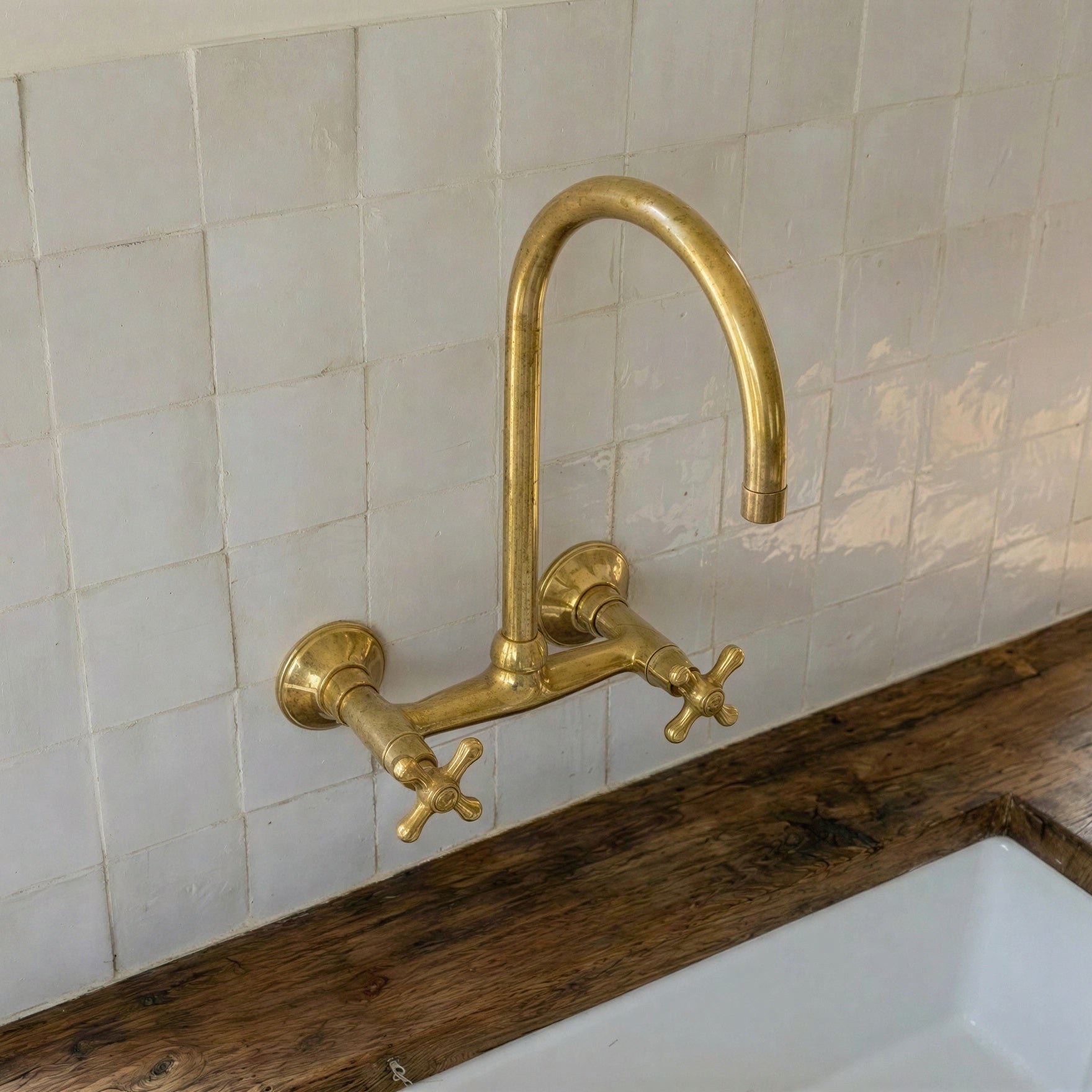 A polished brass wall-mounted kitchen faucet with a high-arc spout and two cross handles, installed above a white sink on a rustic wooden countertop against a white tiled backsplash.