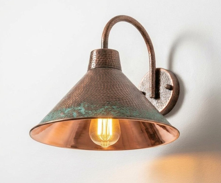 A rustic industrial wall sconce with a hammered copper-colored metal shade and a warm glowing light bulb, mounted on a plain white wall.