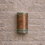 A cylindrical outdoor wall sconce with a distressed bronze finish and green verdigris accents, mounted on a textured stone wall.