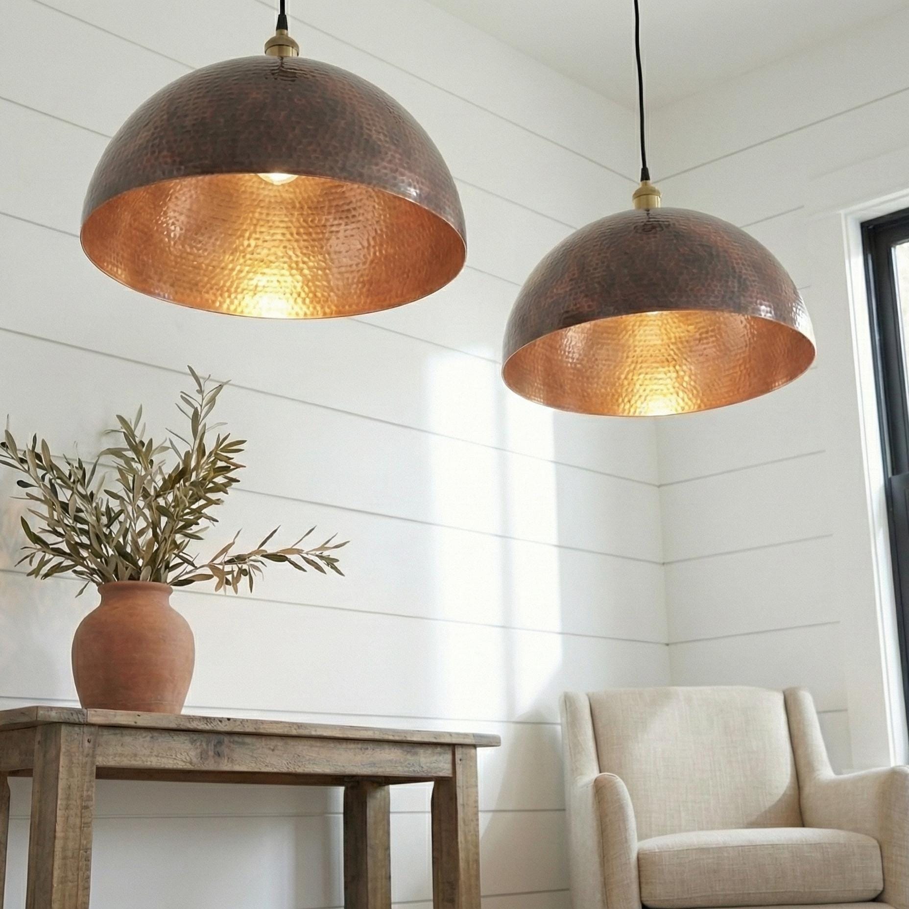 Two copper pendant lights hanging above a table with a plant and chair in a room.