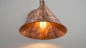 A hammered copper-colored metal pendant light shade glowing with a warm Edison bulb inside, shown against a plain light gray wall.