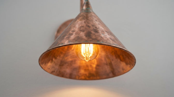 A hammered copper-colored metal pendant light shade glowing with a warm Edison bulb inside, shown against a plain light gray wall.