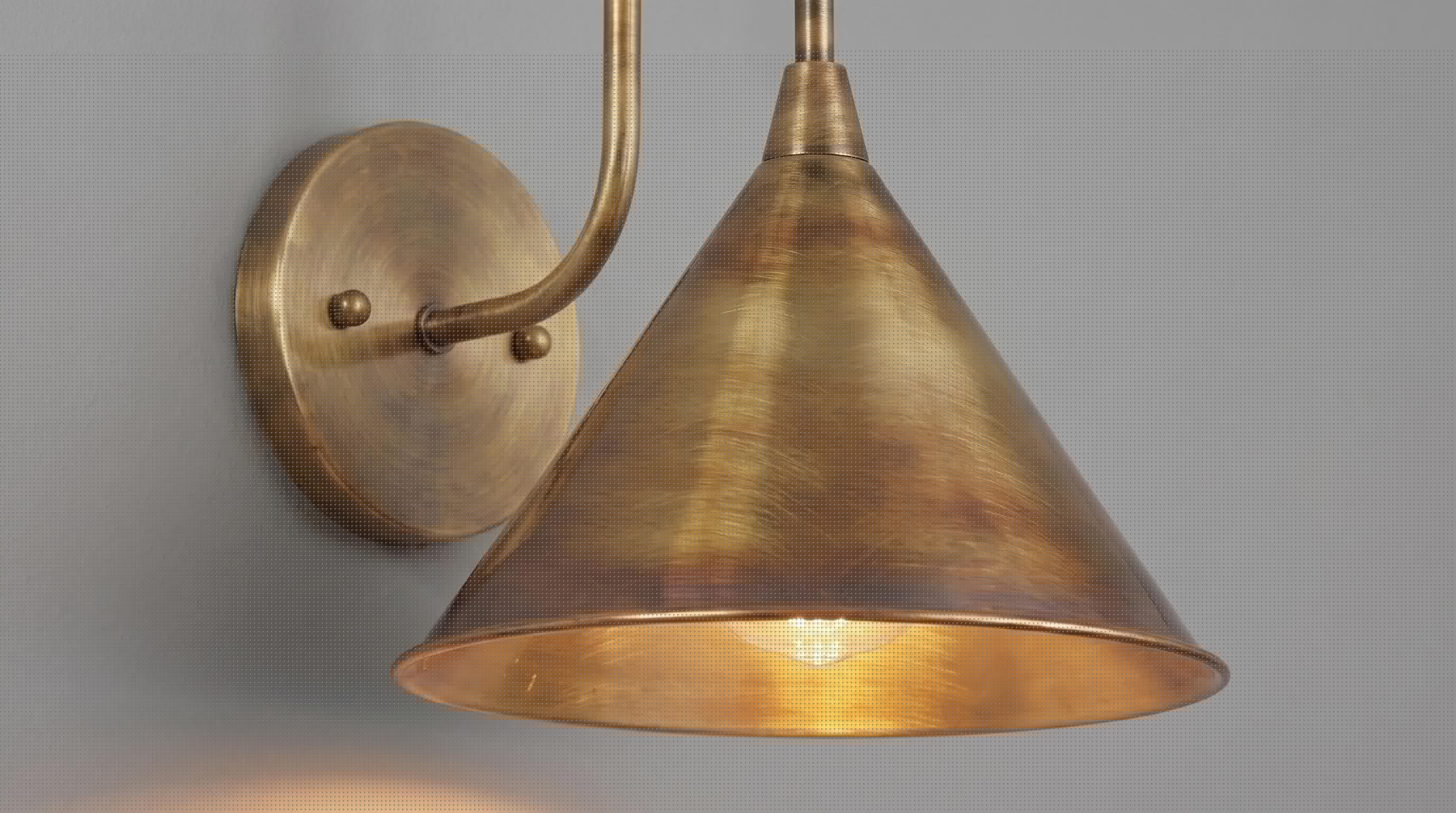 A close-up of a wall-mounted sconce with a warm brass or bronze metal shade and matching wall plate, shown against a plain light gray wall.