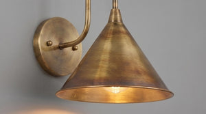 A close-up of a wall-mounted sconce with a warm brass or bronze metal shade and matching wall plate, shown against a plain light gray wall.
