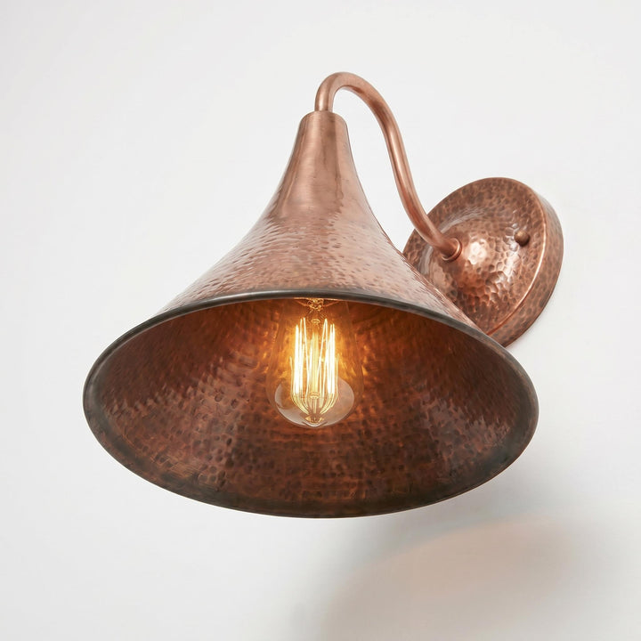 A hammered copper wall sconce with a large conical shade, curved arm, and round wall mount, shown lit against a plain light background.