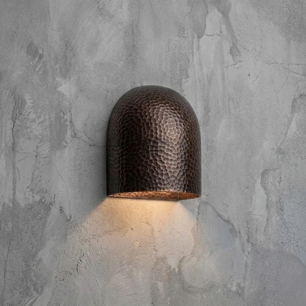 A dome-shaped wall sconce with a hammered metallic finish, mounted on a textured gray concrete wall and casting a warm light downward.