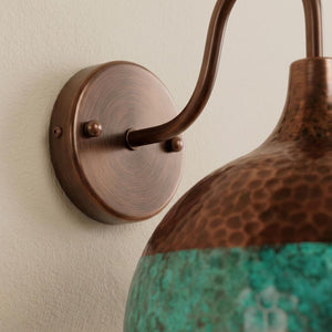 Close-up of a wall-mounted sconce with a round bronze backplate, curved bronze arm, and a hammered copper-toned shade transitioning into a textured turquoise lower section, mounted on a light beige wall.
