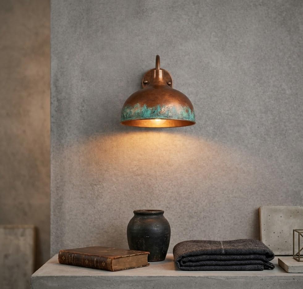 A copper dome wall sconce with a verdigris patina and warm glowing light, mounted on a textured gray concrete wall above a stone ledge with a book, ceramic pot, and folded dark blanket.