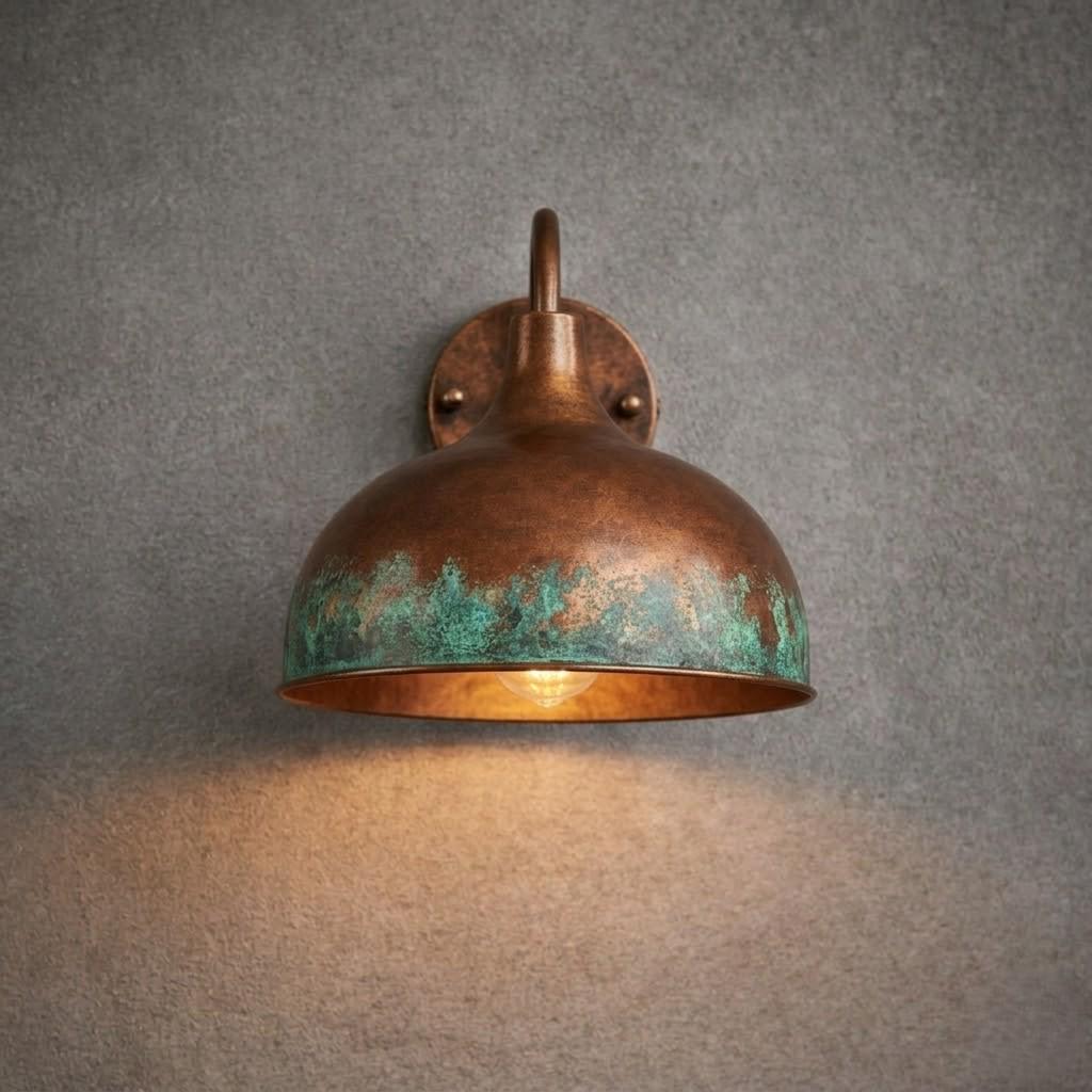 A dome-shaped wall sconce with a distressed bronze finish and a green patina around the lower half, shown illuminated against a textured gray wall.