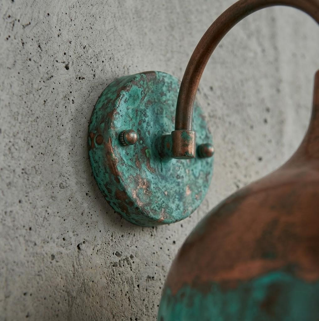 Close-up of a distressed copper wall sconce with a turquoise patina circular backplate mounted on a textured concrete wall.