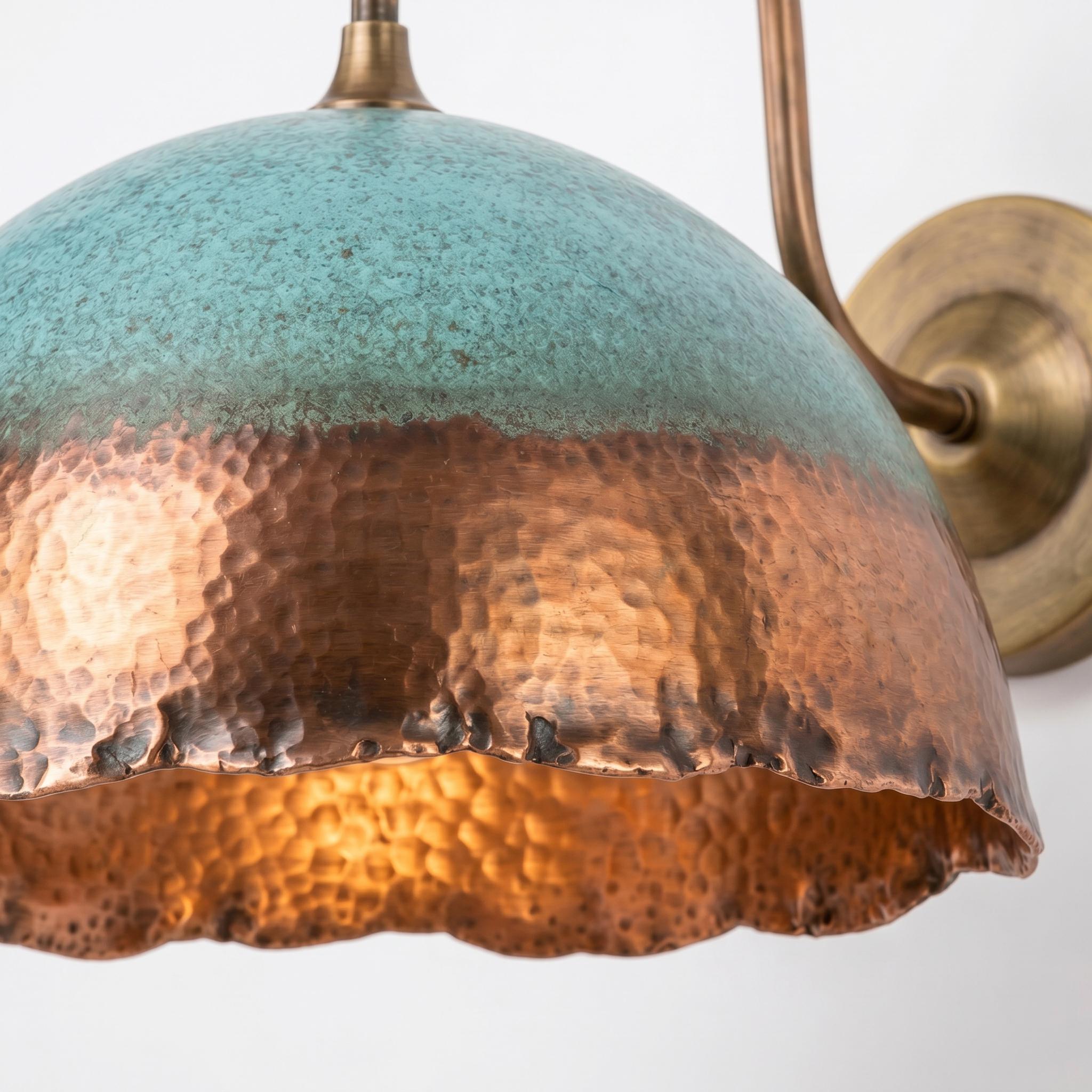 A close-up of a hammered metal dome pendant light shade with a turquoise-blue top half and a copper-toned bottom half, shown against a plain light background with part of the wall mount visible.