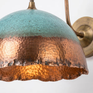 A close-up of a hammered metal dome pendant light shade with a turquoise-blue top half and a copper-toned bottom half, shown against a plain light background with part of the wall mount visible.