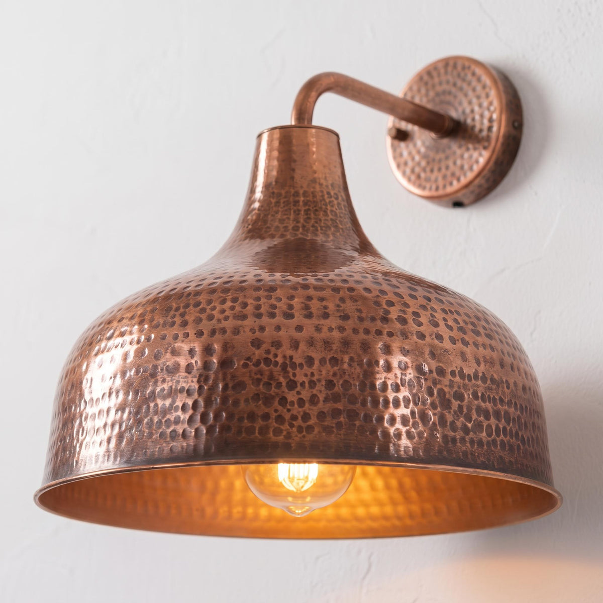 A hammered copper dome wall sconce with a warm glowing light bulb inside, mounted on a textured white wall.
