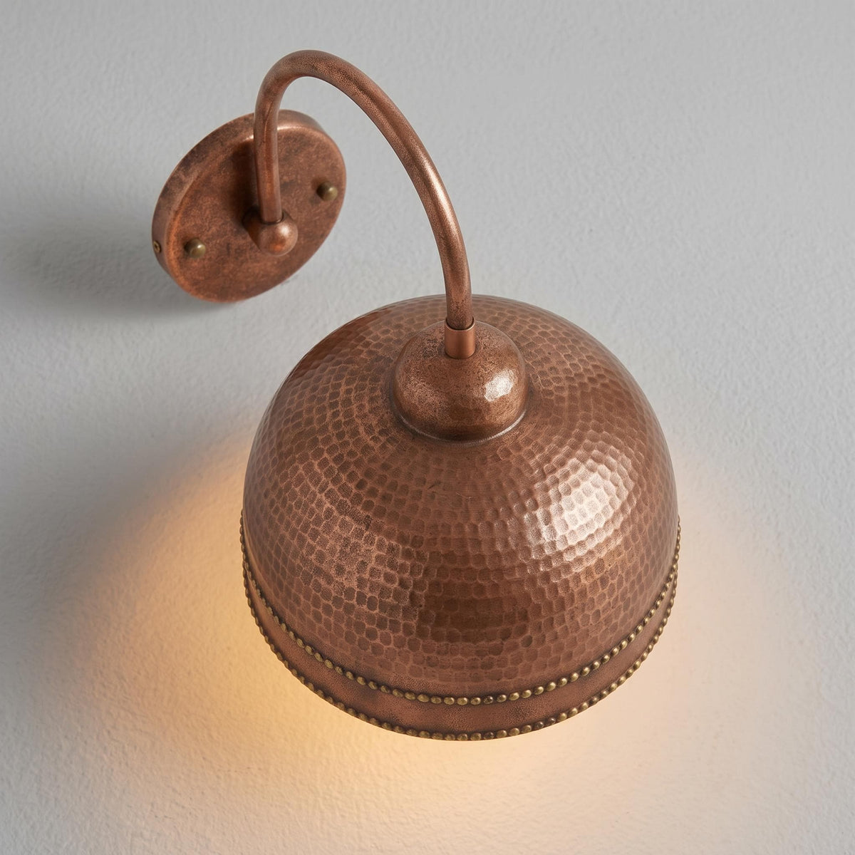 A hammered copper dome wall sconce with a curved arm and round mounting plate, shown lit against a light gray wall.