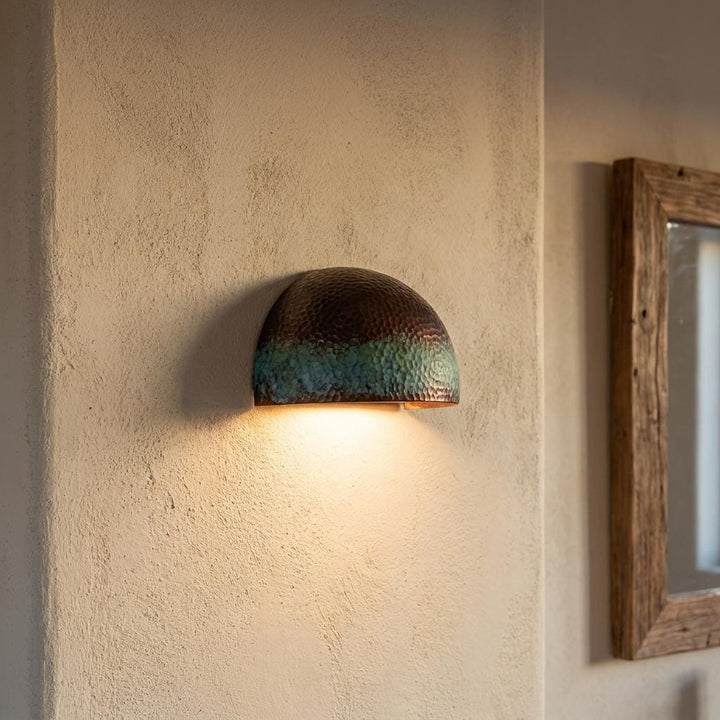A dome-shaped wall sconce with a textured metallic finish, featuring a dark bronze top half and a green-blue lower half, mounted on a textured beige plaster wall and casting a warm light downward.