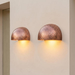 Two hammered copper dome wall sconces mounted side-by-side on a textured beige stucco wall, casting a warm yellow light downward.